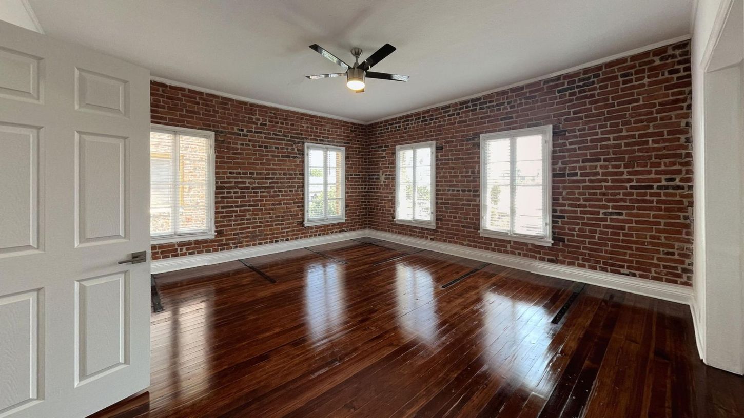 Empty room with brick walls, hardwood floors, and multiple windows. A door is on the left.