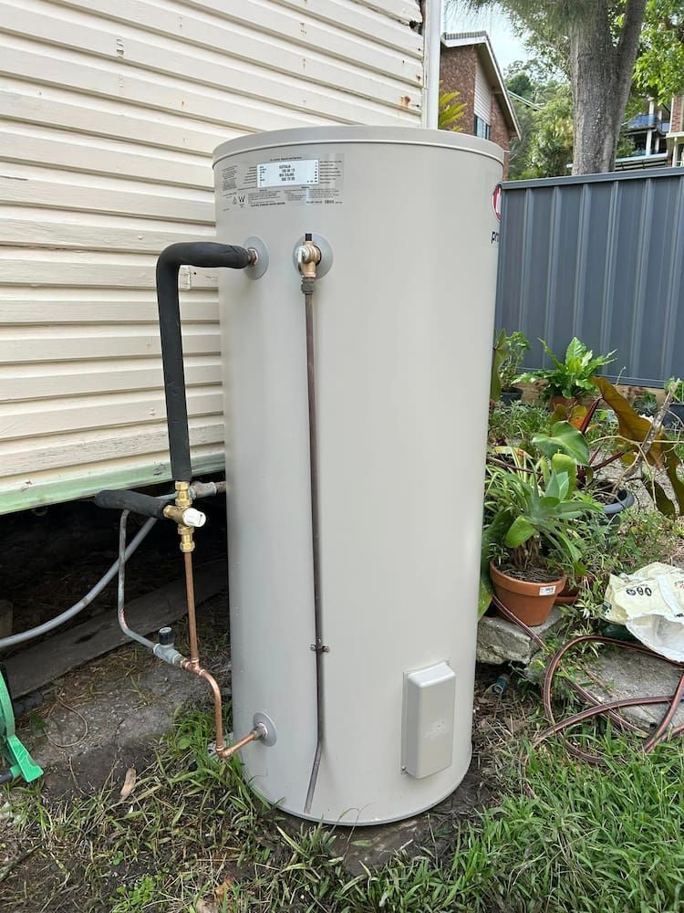 A Large White Water Heater Is Sitting In The Grass Next To A House — DMK Plumbing in Crescent Head, NSW