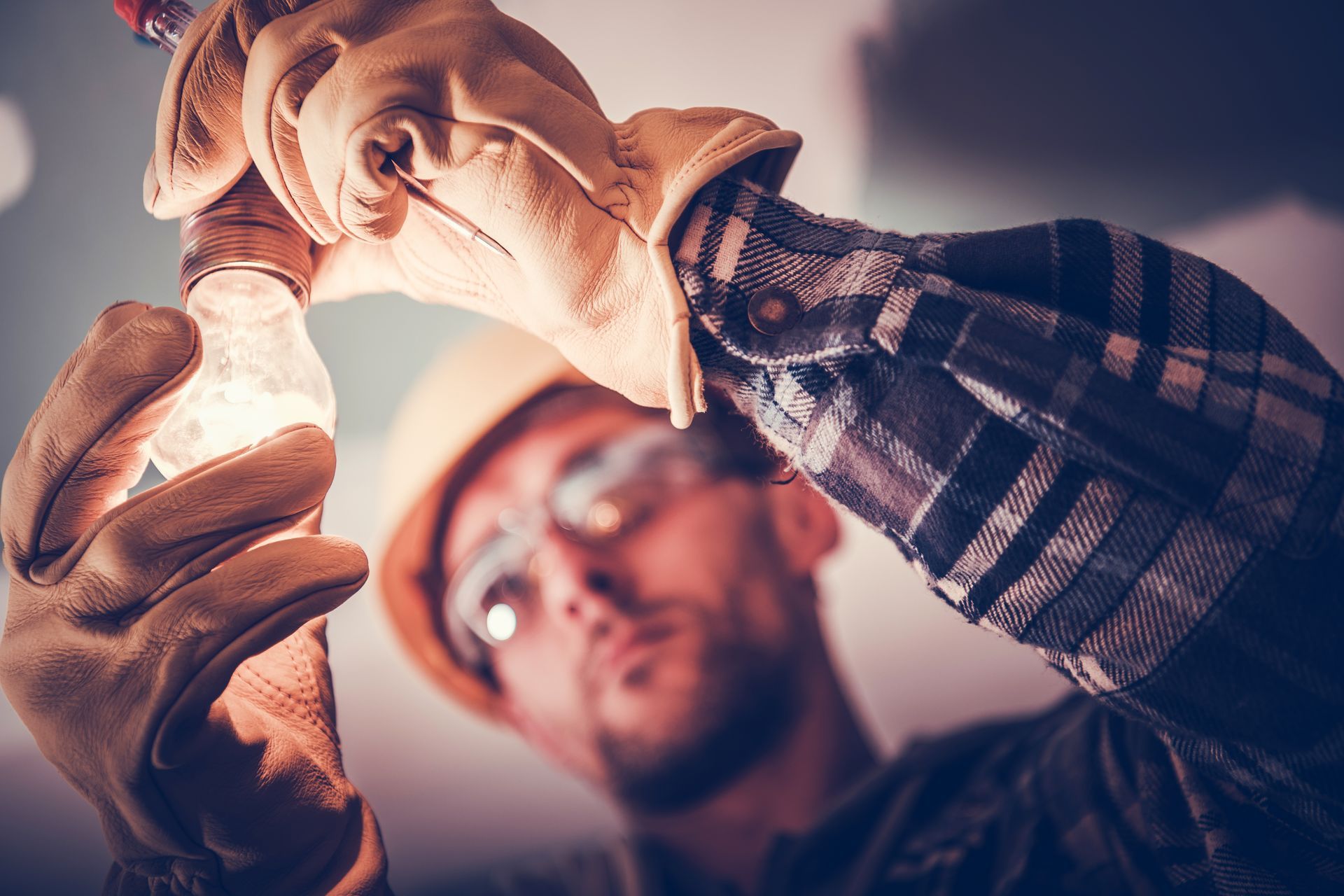 Electrician replacing a lightbulb, wearing gloves, safety glasses, and a hard hat.
