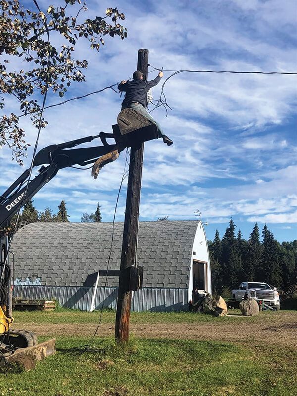 Lineman atop a utility pole repairs wires with a lift bucket on a sunny day. A white barn is in the background.