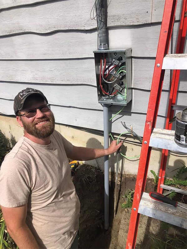 Man points to an electrical box installed on a gray pole next to a house; red ladder is nearby.
