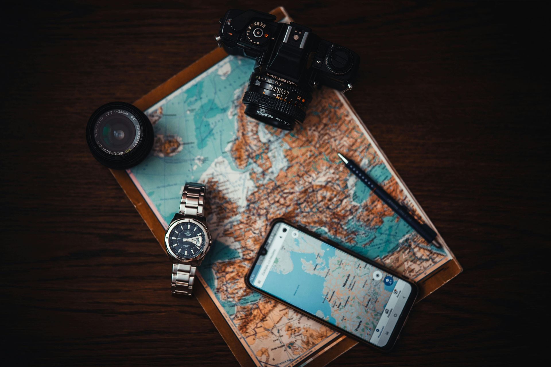 Travel gear on a wooden table: folded map, smartphone, camera, watch, and compass.
