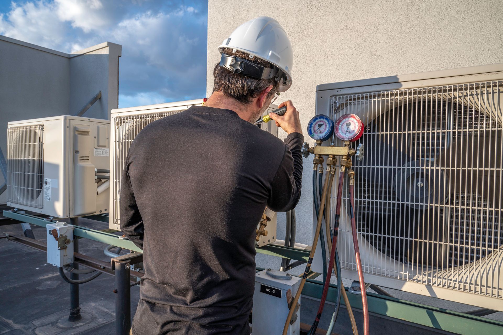HVAC technician in a hard hat repairs rooftop air conditioning unit, using gauges.