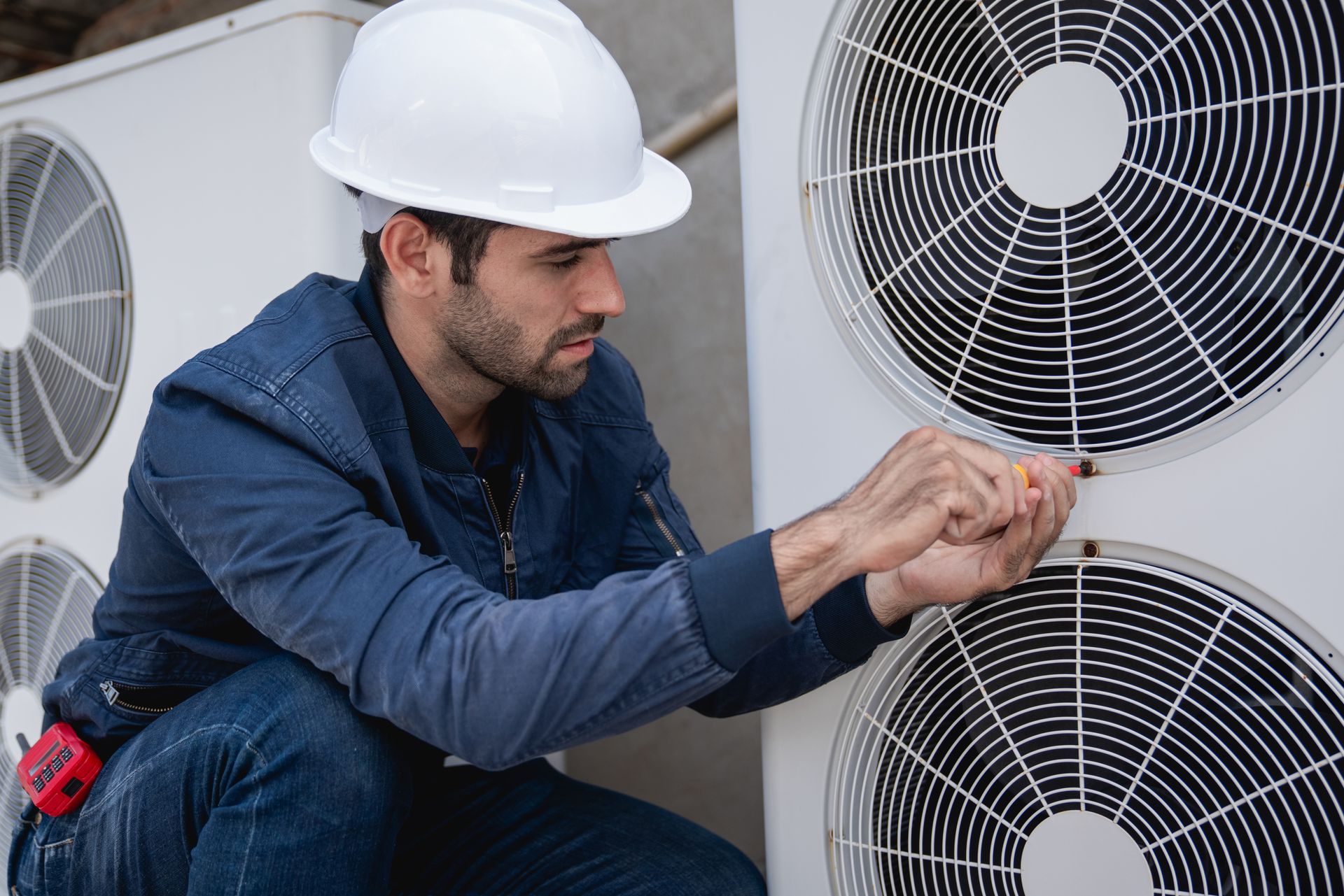 Man in hard hat repairs an outdoor HVAC unit, crouching, using a screwdriver.