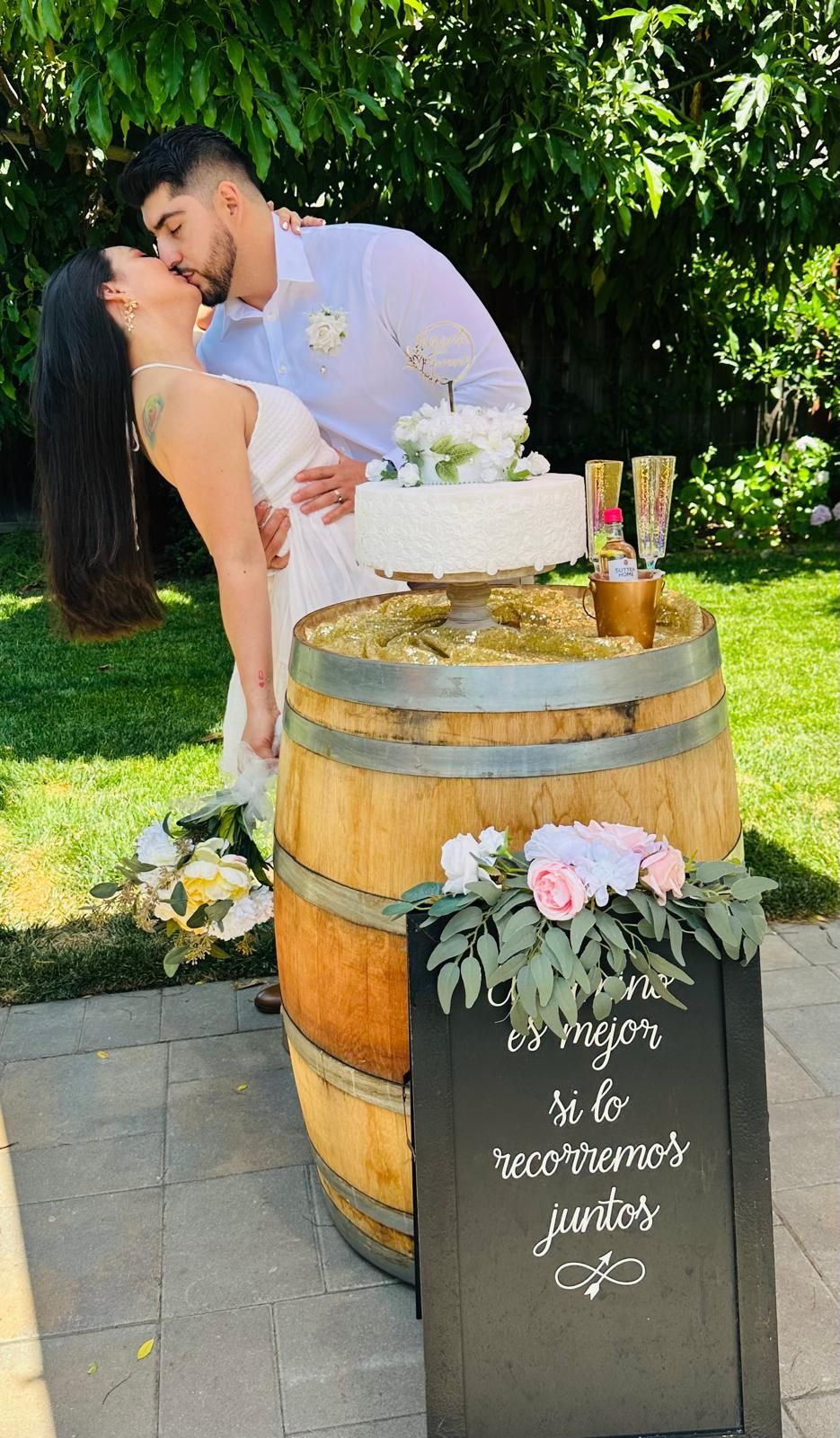 Couple kissing by a barrel cake stand at outdoor wedding; white dress, cake, flowers, Bodas by Lina, Santa Clara, CA.