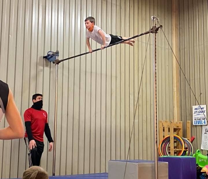 A young boy performs a suspended plank exercise on a bar, supervised by an instructor in a gym setting.