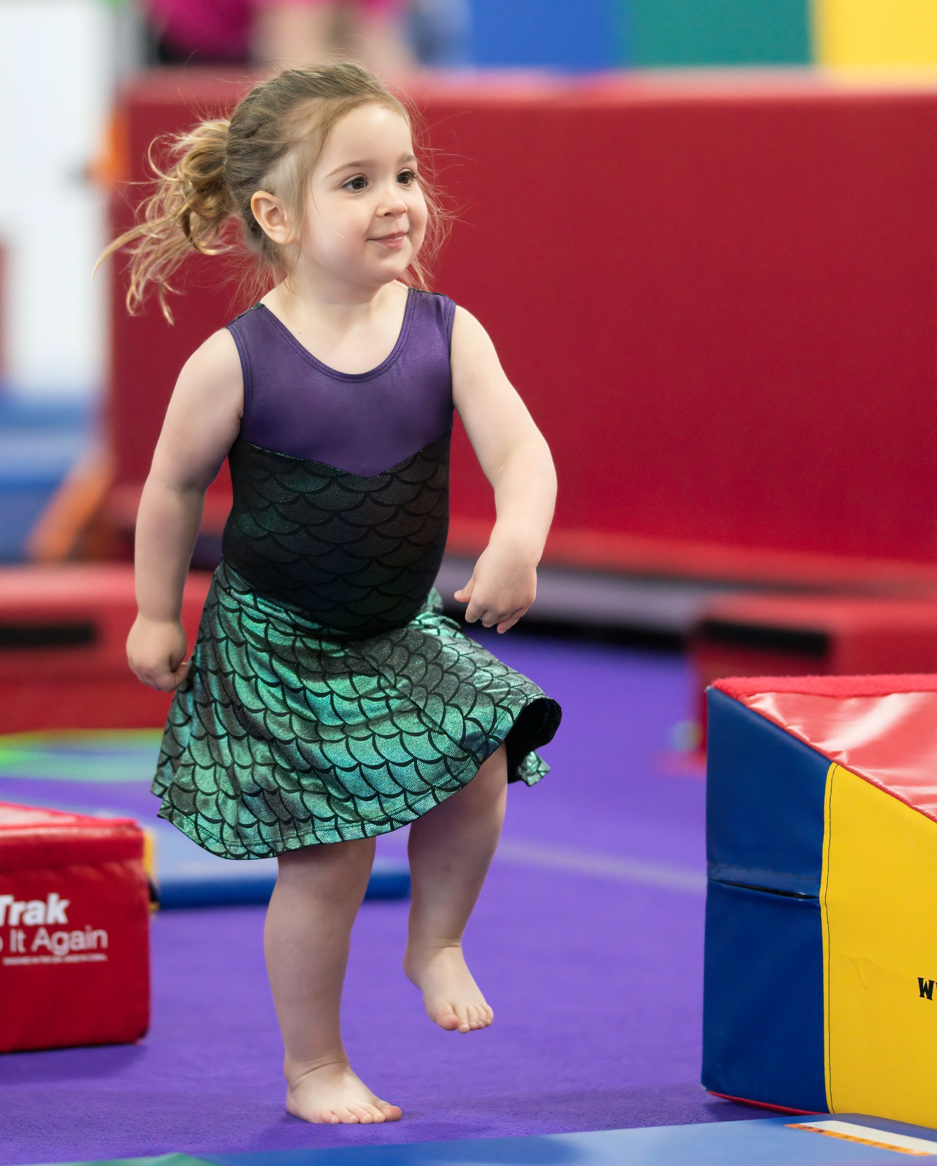 A young girl in a mermaid-themed leotard and skirt smiles as she steps forward on a gymnastics mat.