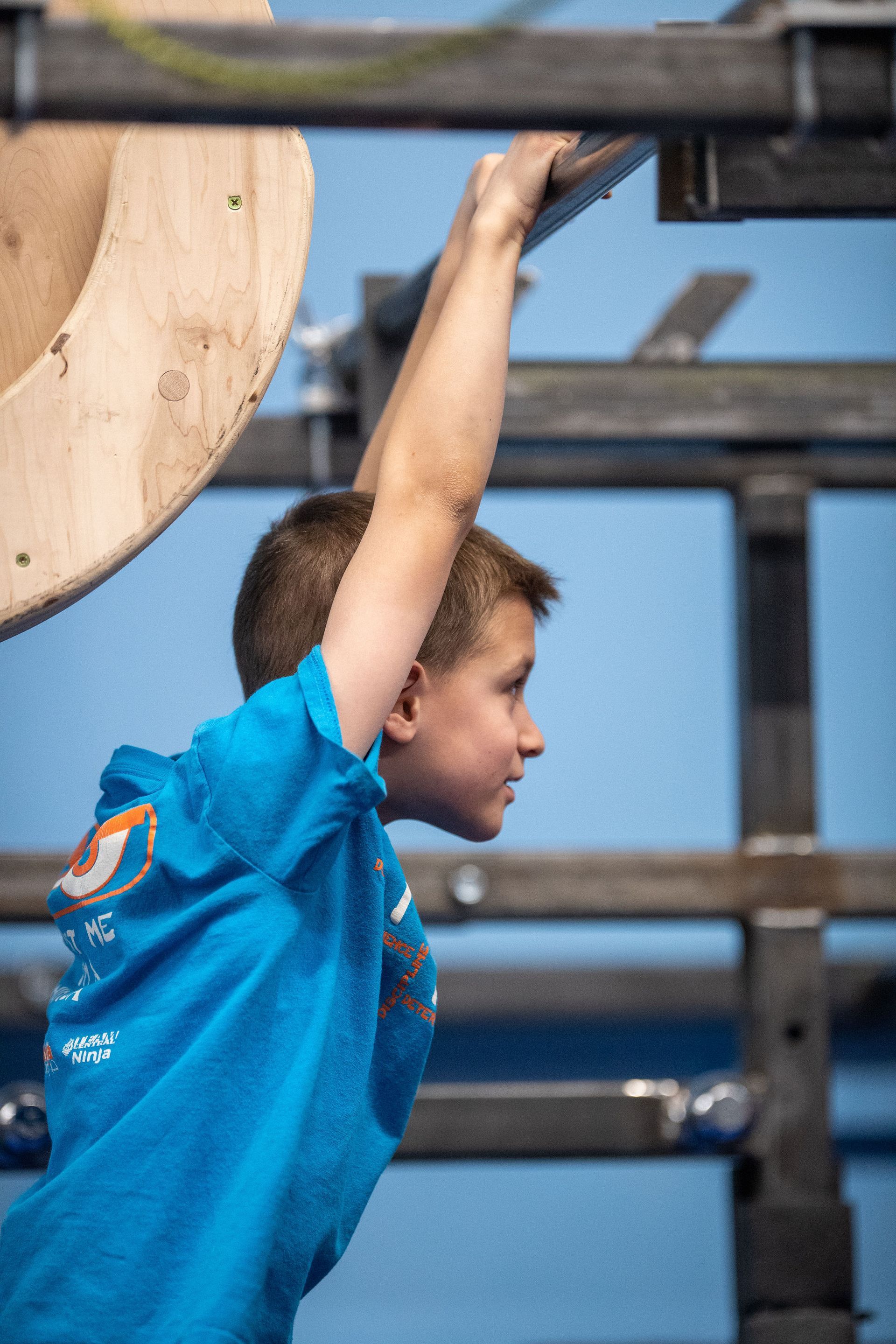 Young boy in a blue shirt grips a climbing structure. He is focused, with his arm extended.