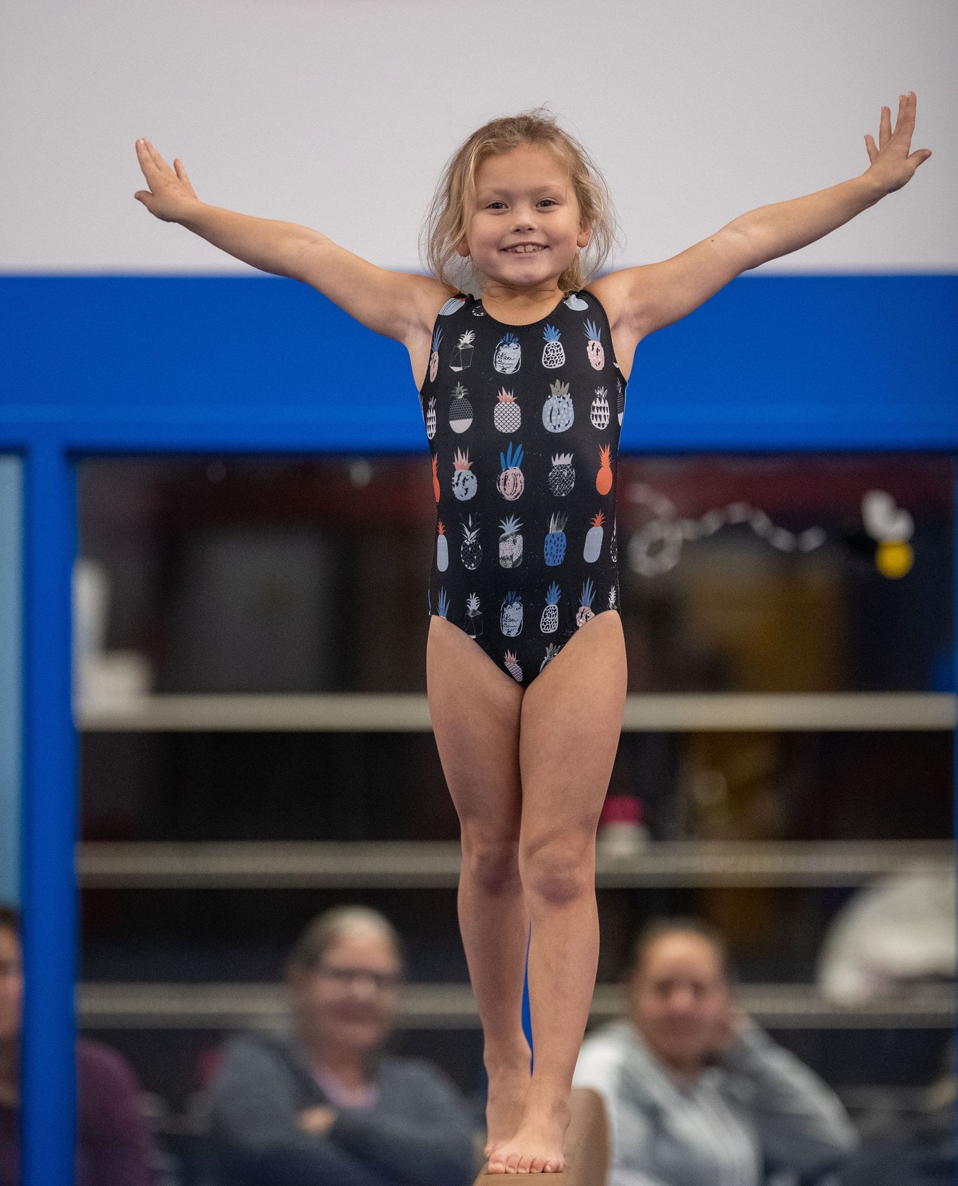 A young girl balances on a beam, arms outstretched, smiling. She wears a black leotard with a cartoon pattern.
