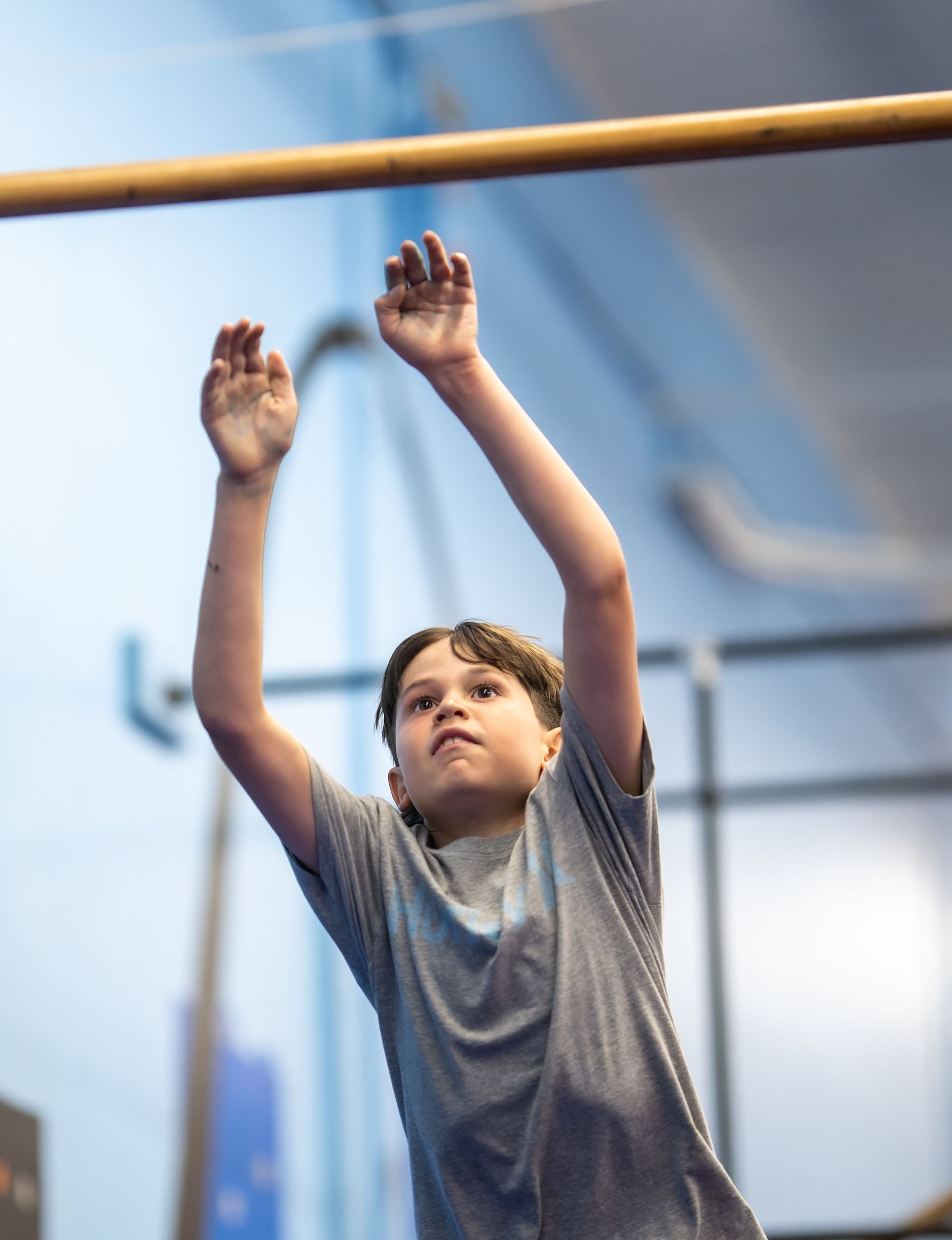 Young boy reaching for a bar, likely in a gym. He wears a gray shirt and has a focused expression.