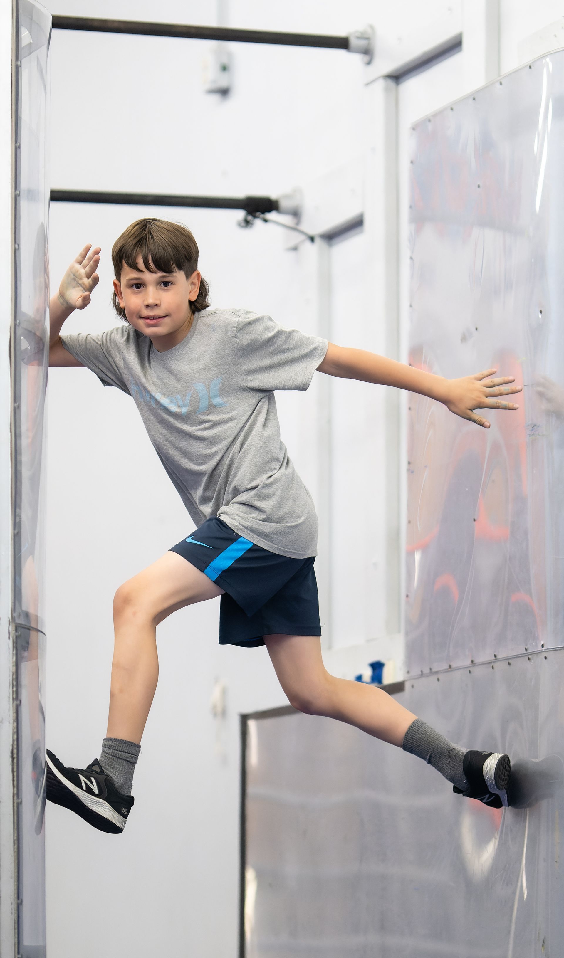 A young boy in athletic wear performs a climbing exercise on a metal wall, reaching and balancing with a smile.