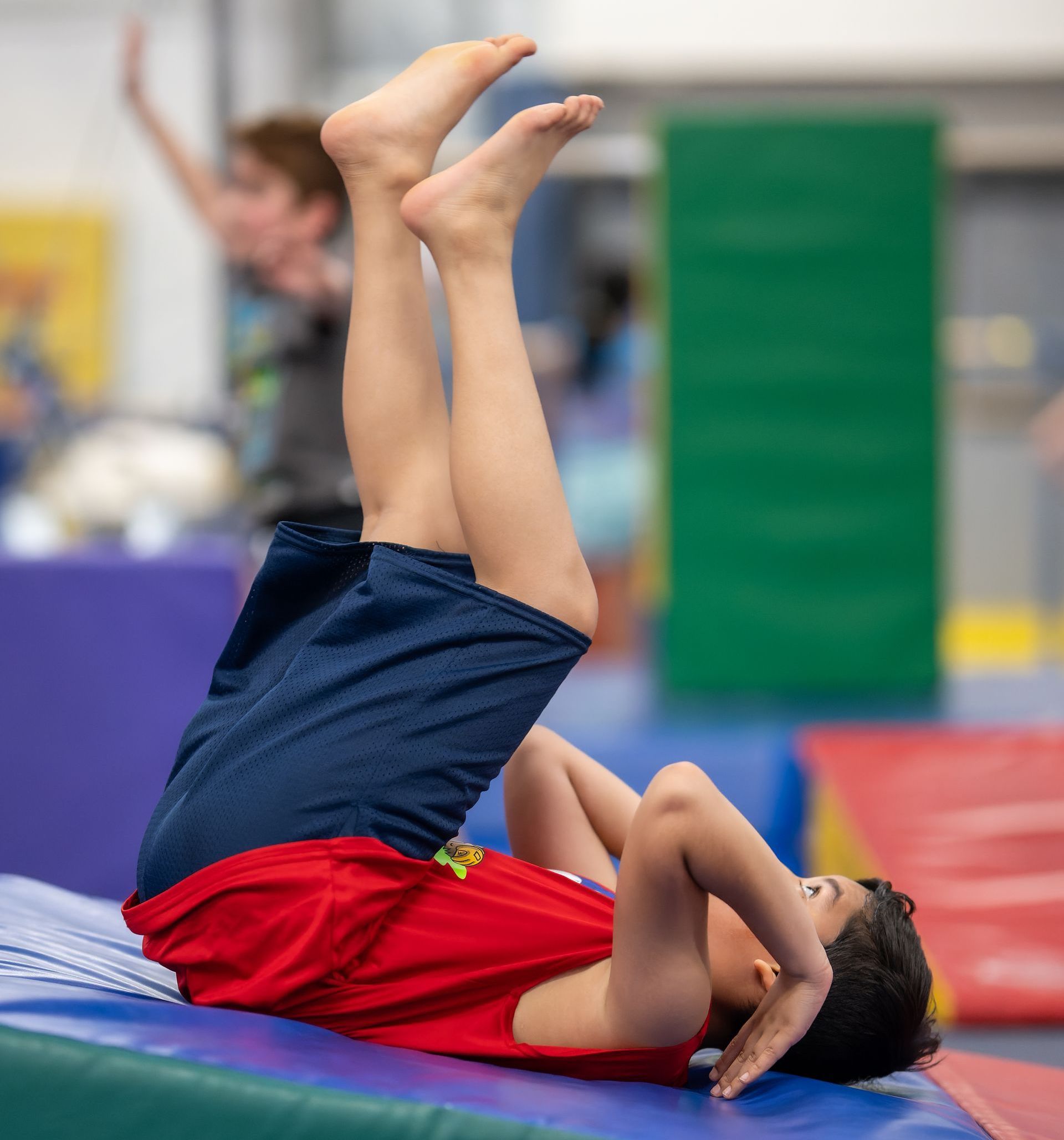 Boy in red shirt and blue shorts lying on his back on a mat, legs raised, hands near head. A second boy is in the background.