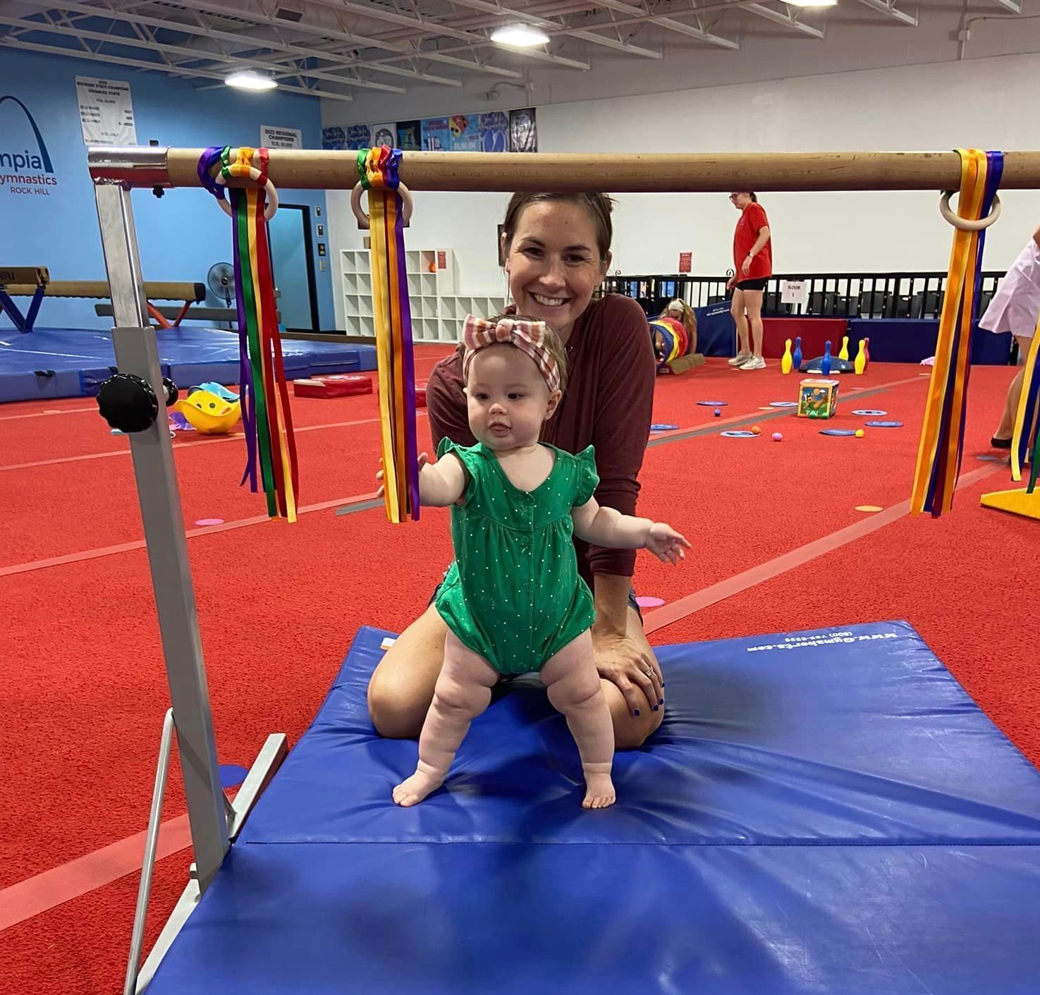 Woman and baby in green romper standing near a bar in a gym.