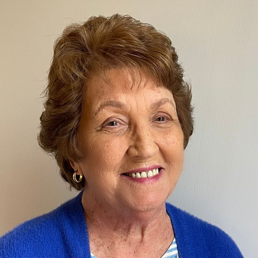 An older woman with short brown hair smiles at the camera, wearing a blue sweater and gold hoop earrings.