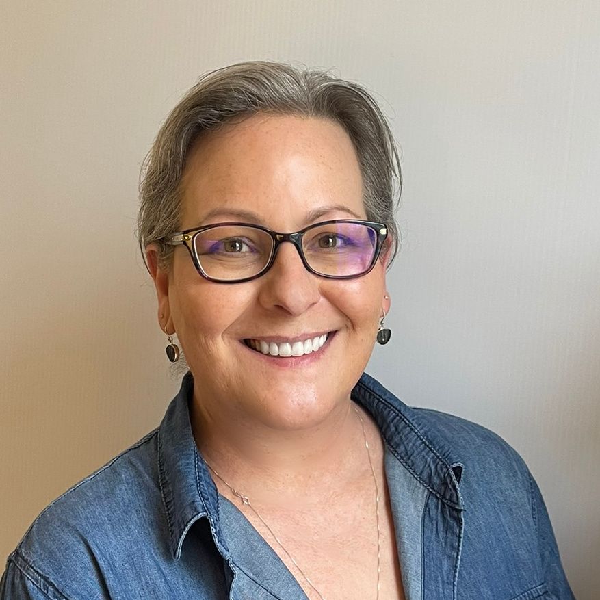 A woman with short gray hair and glasses smiles at the camera, wearing a blue denim shirt.