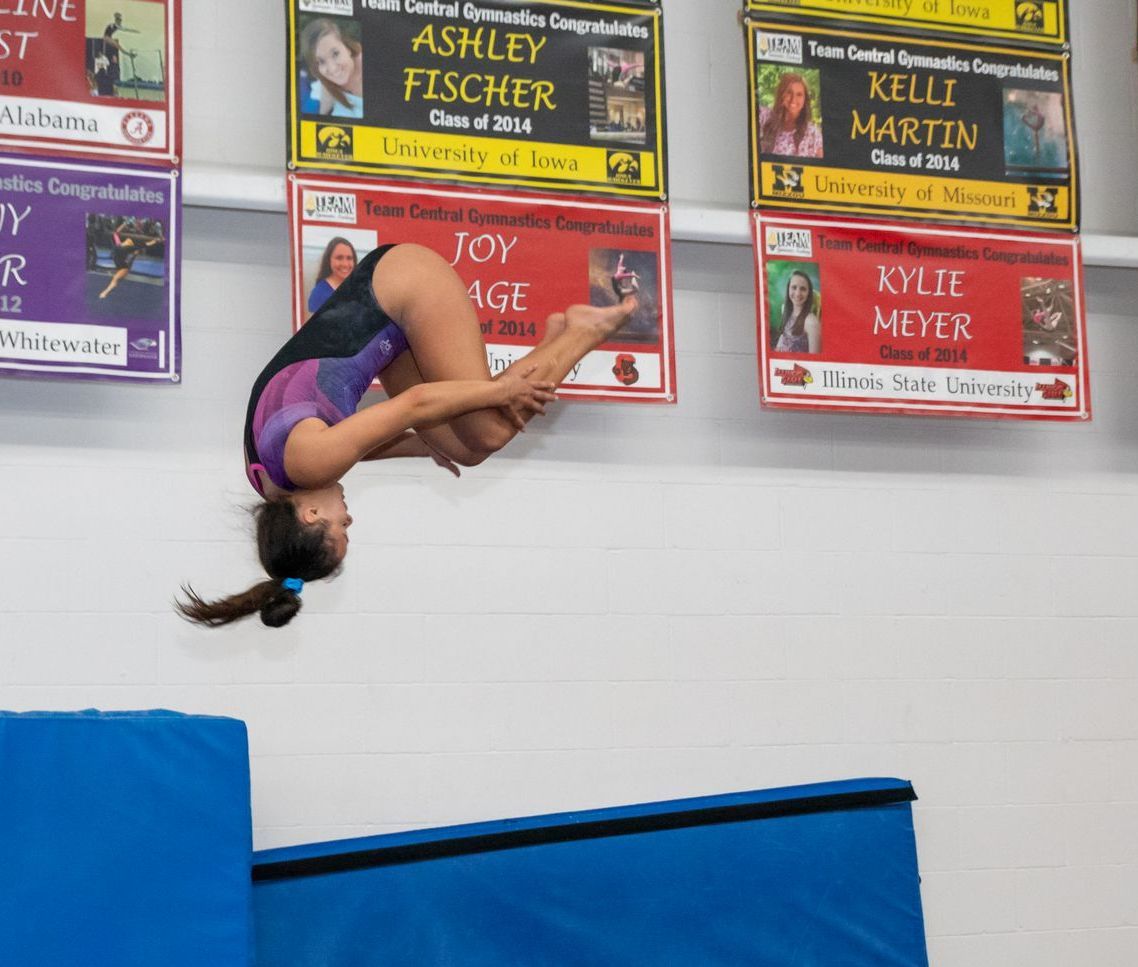 Gymnast in mid-air backflip over a blue mat, performing in a gym.