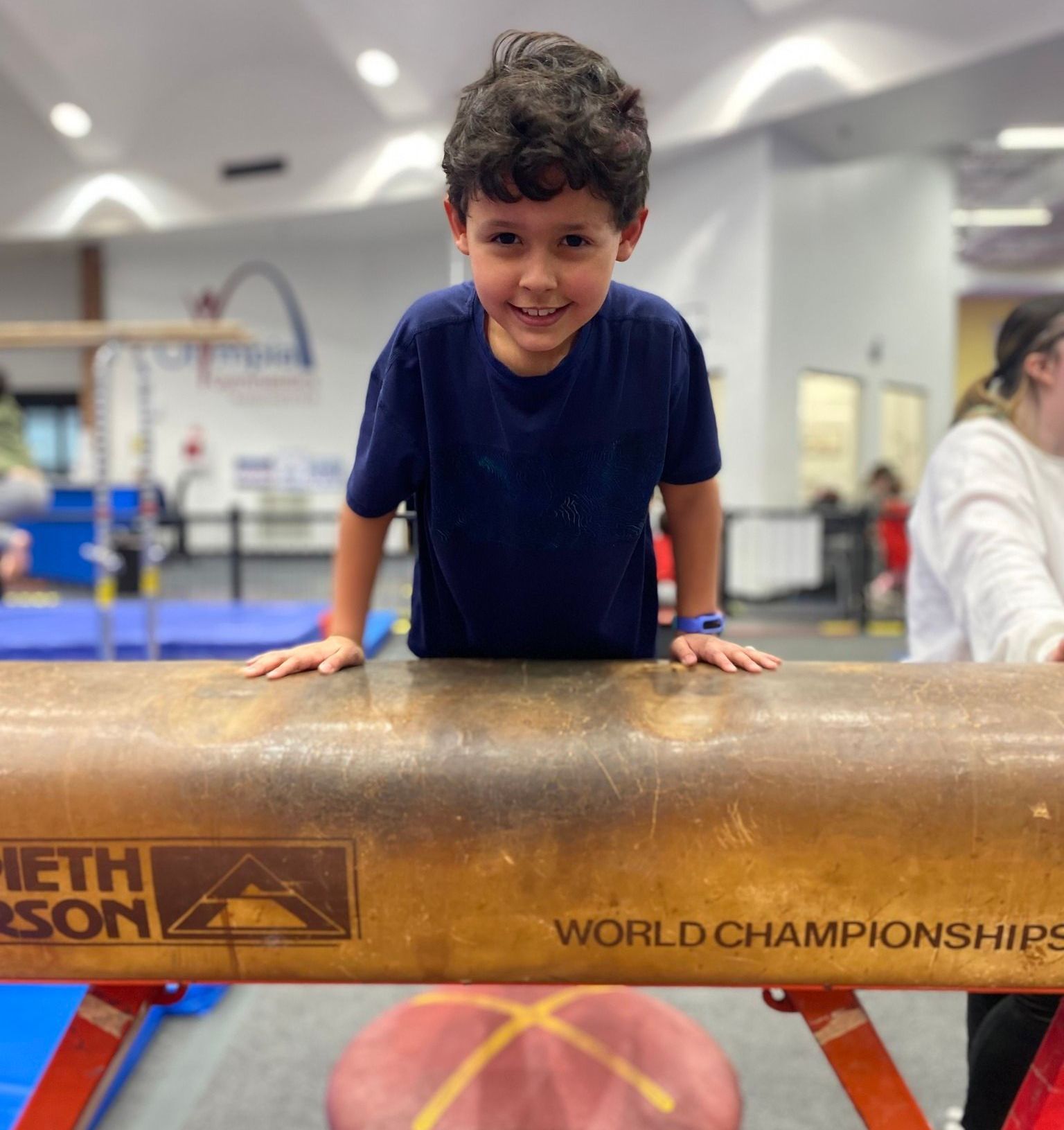 Young boy with dark hair smiles, standing on a gymnastics pommel horse, in a gym with equipment.