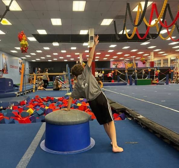 A person doing a gymnastics skill, balancing on a blue cylinder in a gym. They are wearing a grey shirt and black shorts.