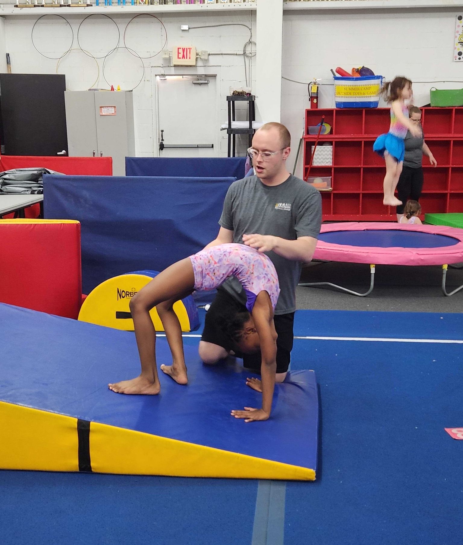 A young girl doing a bridge pose, with a coach assisting in a gymnastics gym. The girl wears a leotard; coach kneels.