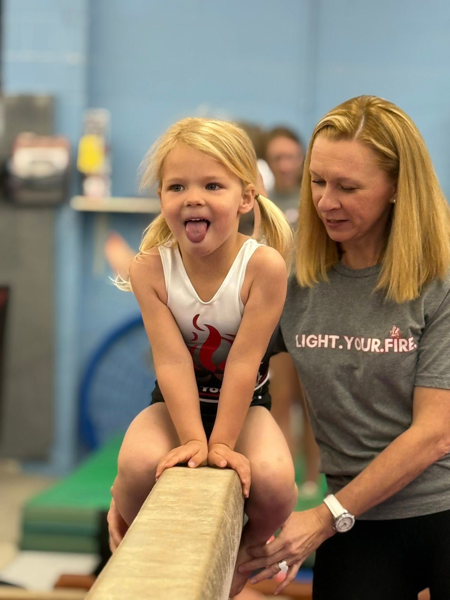 A young girl with pigtails sticks her tongue out while sitting on a balance beam, watched by a coach in a gym.