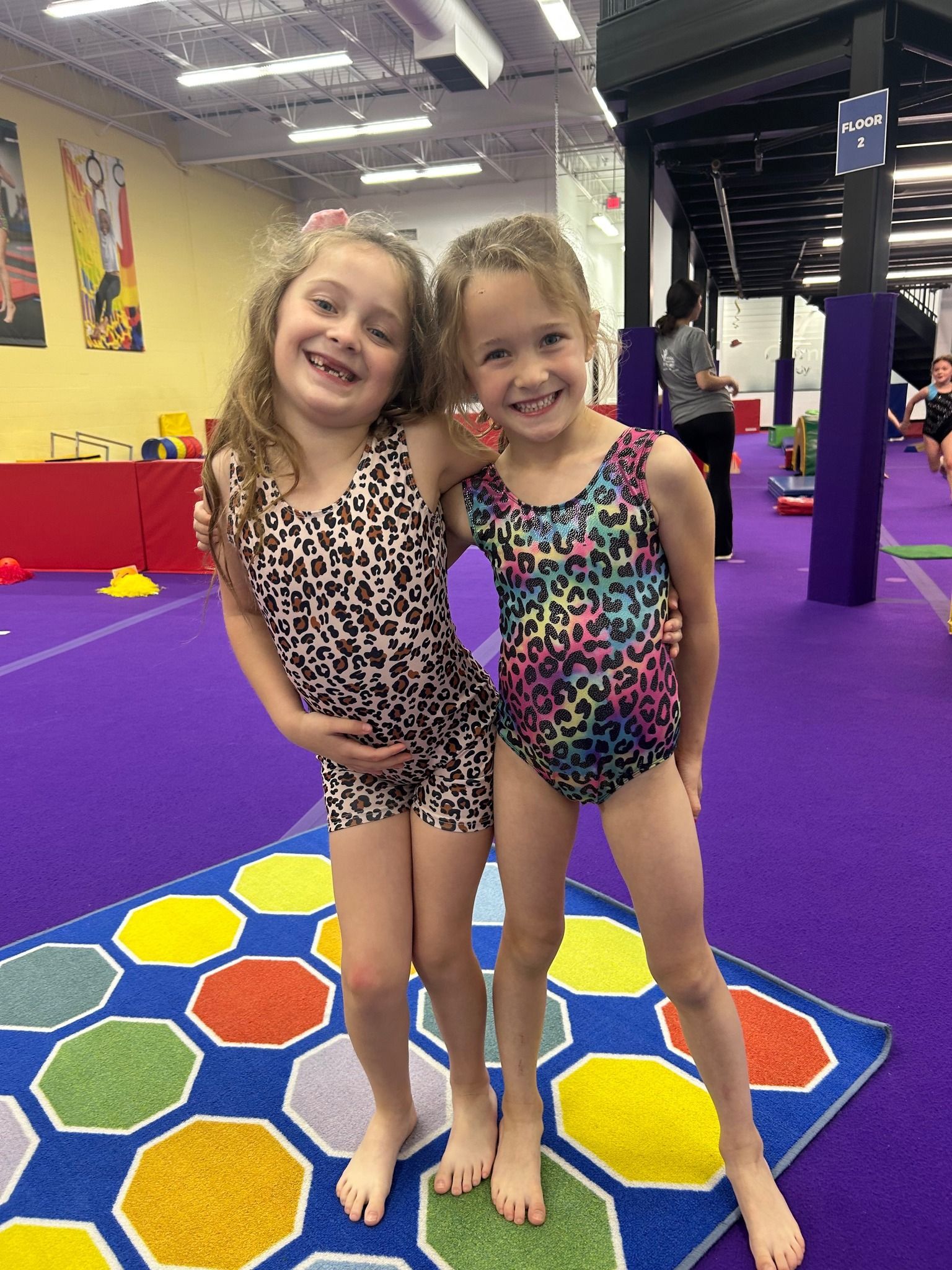 Two young girls in leotards smile and pose together on a gym floor, one with a leopard print leotard.