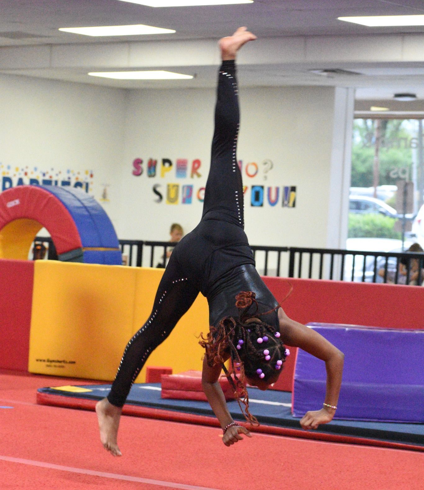 A young girl in black leotard and leggings does a handstand split on a red mat in a gym.