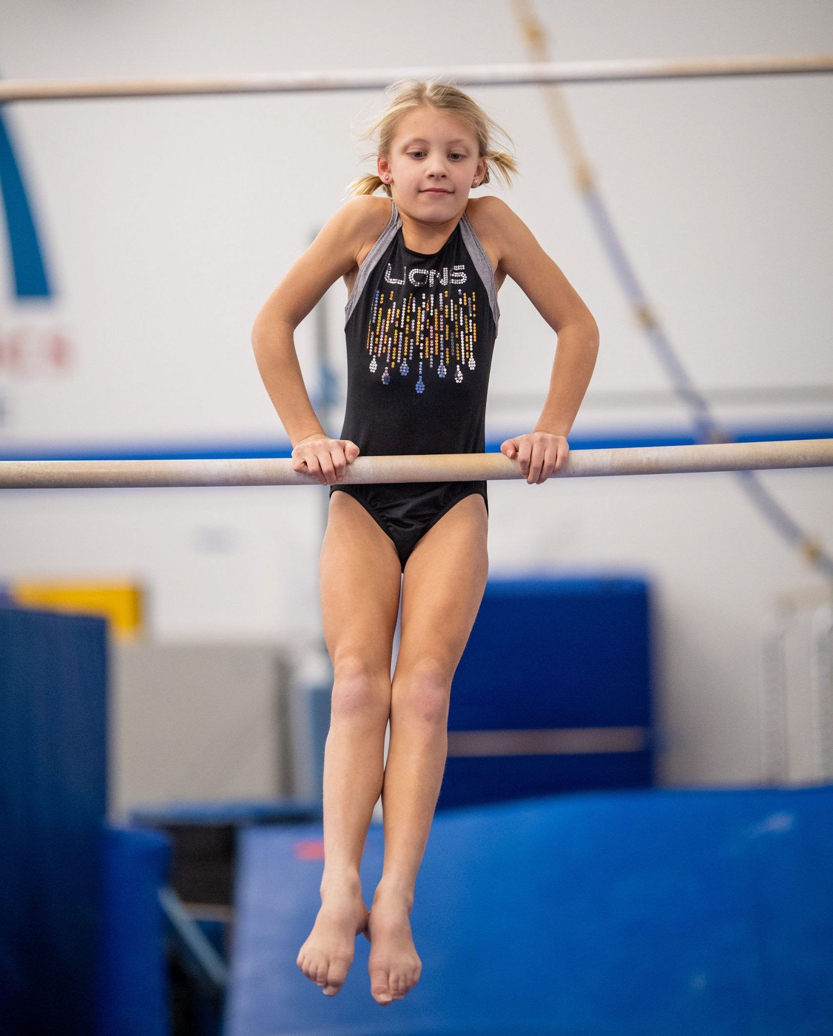 Young gymnast hangs from a horizontal bar in a gym. She wears a black leotard and looks forward.