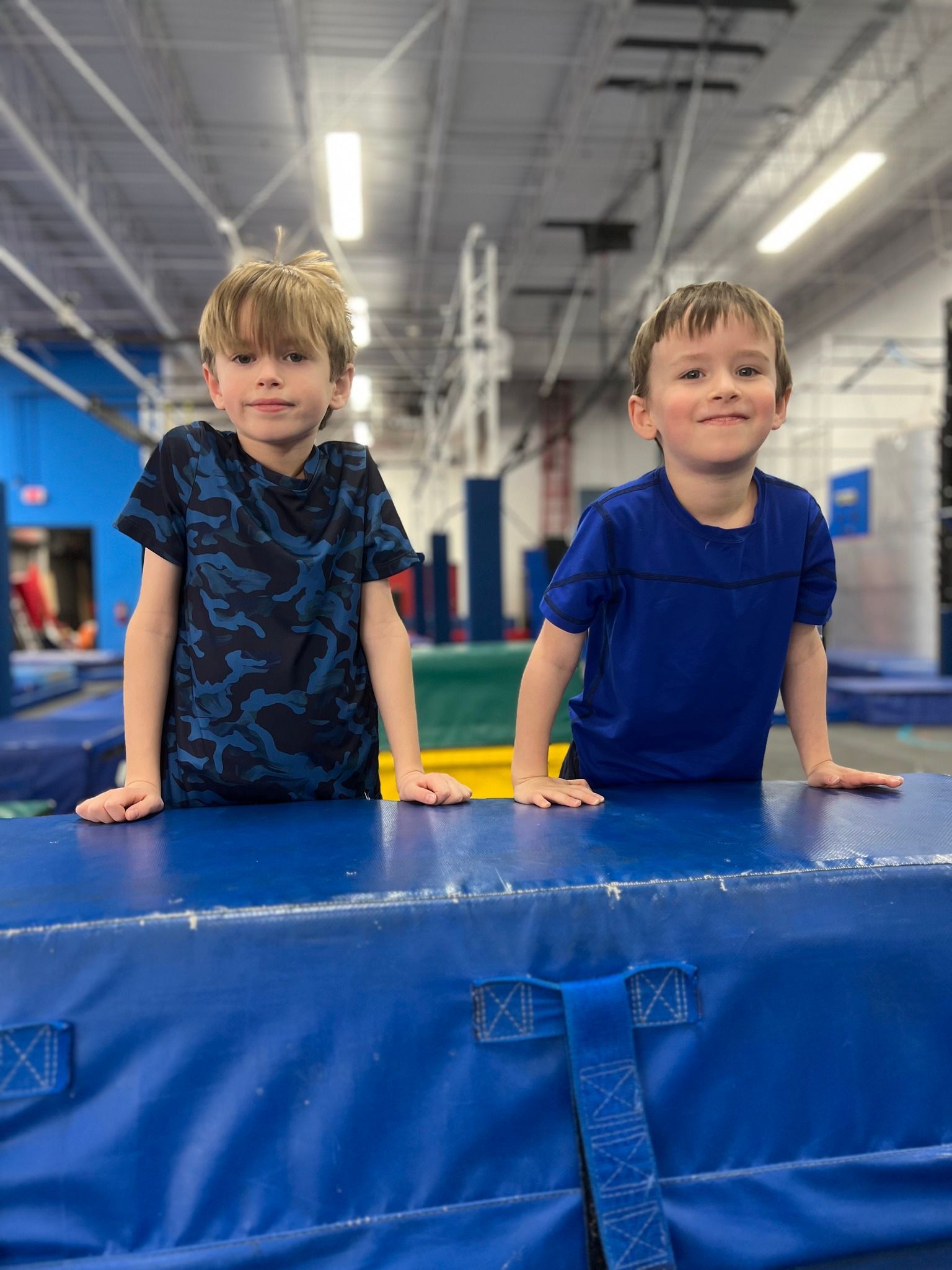 Two young boys, wearing blue shirts, stand smiling behind a blue padded surface in a gym.