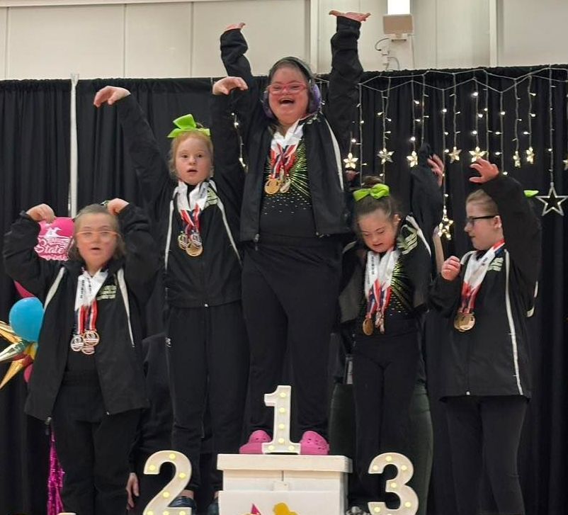 HUGS on a podium, wearing medals, raising arms in celebration. Stage lit with star lights.