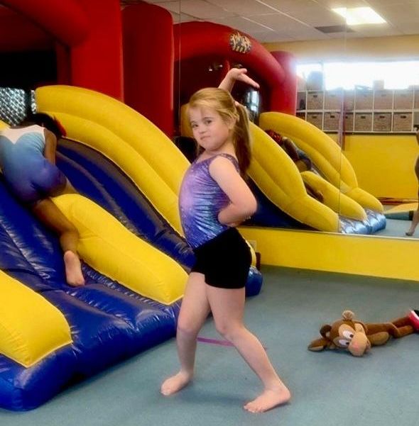 A young girl in a gymnastics leotard poses confidently in a gym with a bouncy slide.