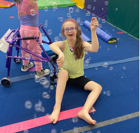 HUGS girl smiling, surrounded by bubbles, sitting on blue gym mat with walker.