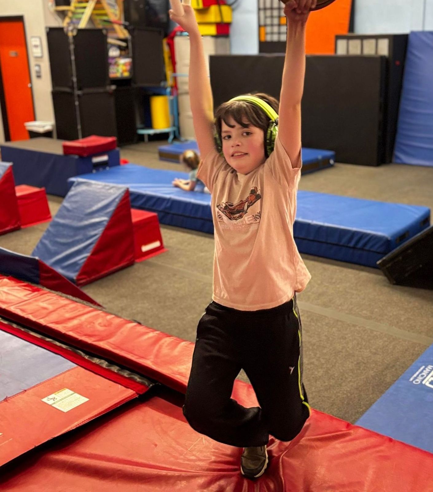 Young boy in black pants and pink shirt hanging from a bar in an indoor obstacle course, arms raised.