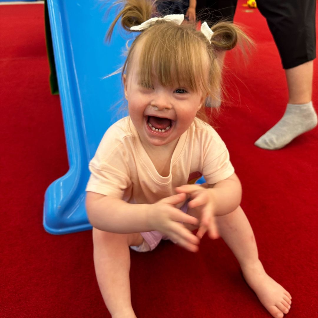 A smiling toddler with pigtails, pink shirt, and bare feet sits on a red surface near a blue slide, clapping.