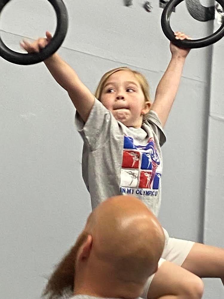 A young girl hanging from gymnastic rings, supported on the shoulders of a man with a shaved head, in a gym.