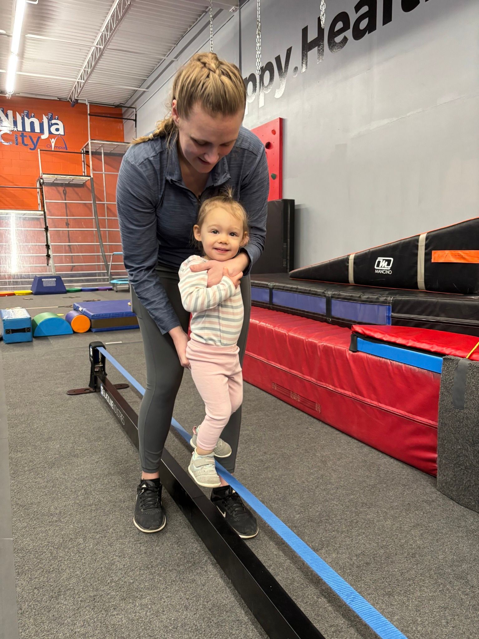 A woman helps a young child walk along a balance beam at a ninja gym. The child smiles as she takes steps.