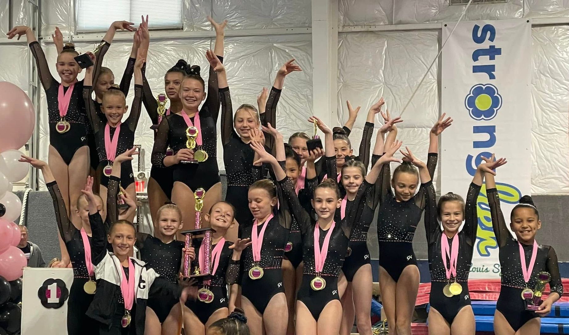 A gymnastics team, wearing black leotards and pink medals, celebrates a victory with arms raised in a gym.