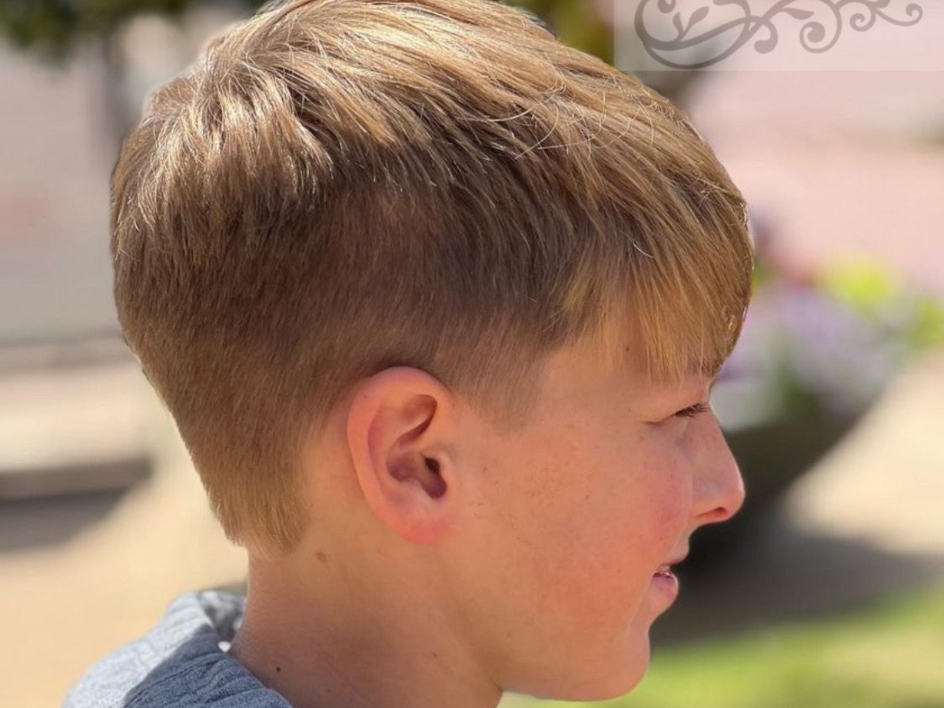 Blond-haired person with a fresh haircut, wearing a gray hoodie, profile view. Outdoors, daytime.