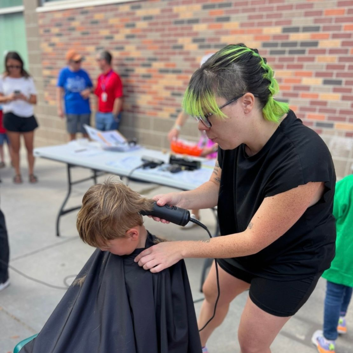 Person with green hair giving a boy a haircut outdoors, near a brick building.