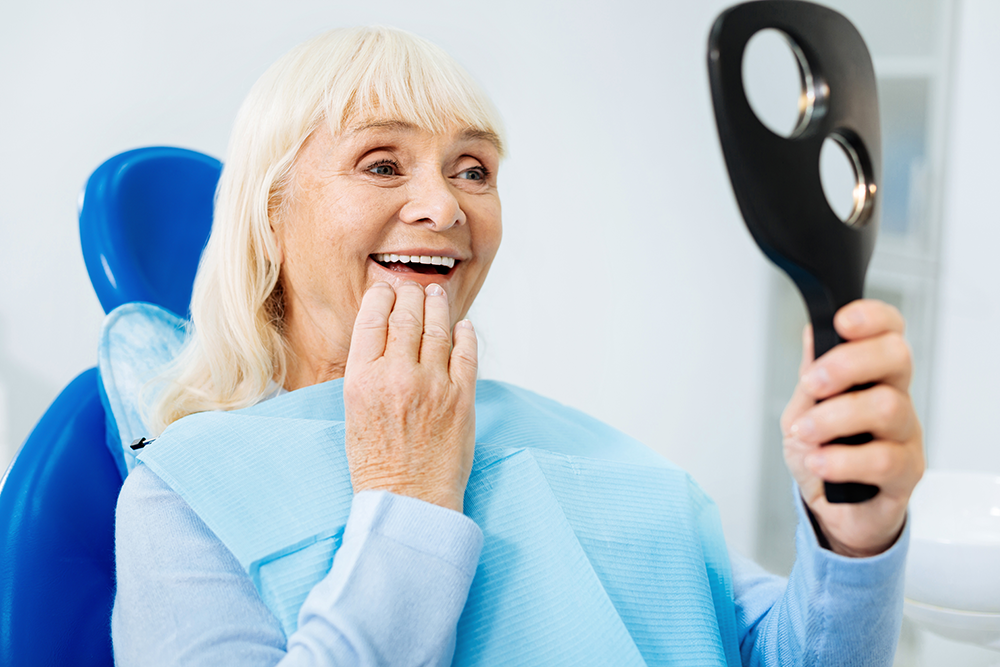 An elderly woman is sitting in a dental chair looking at her teeth in a mirror.