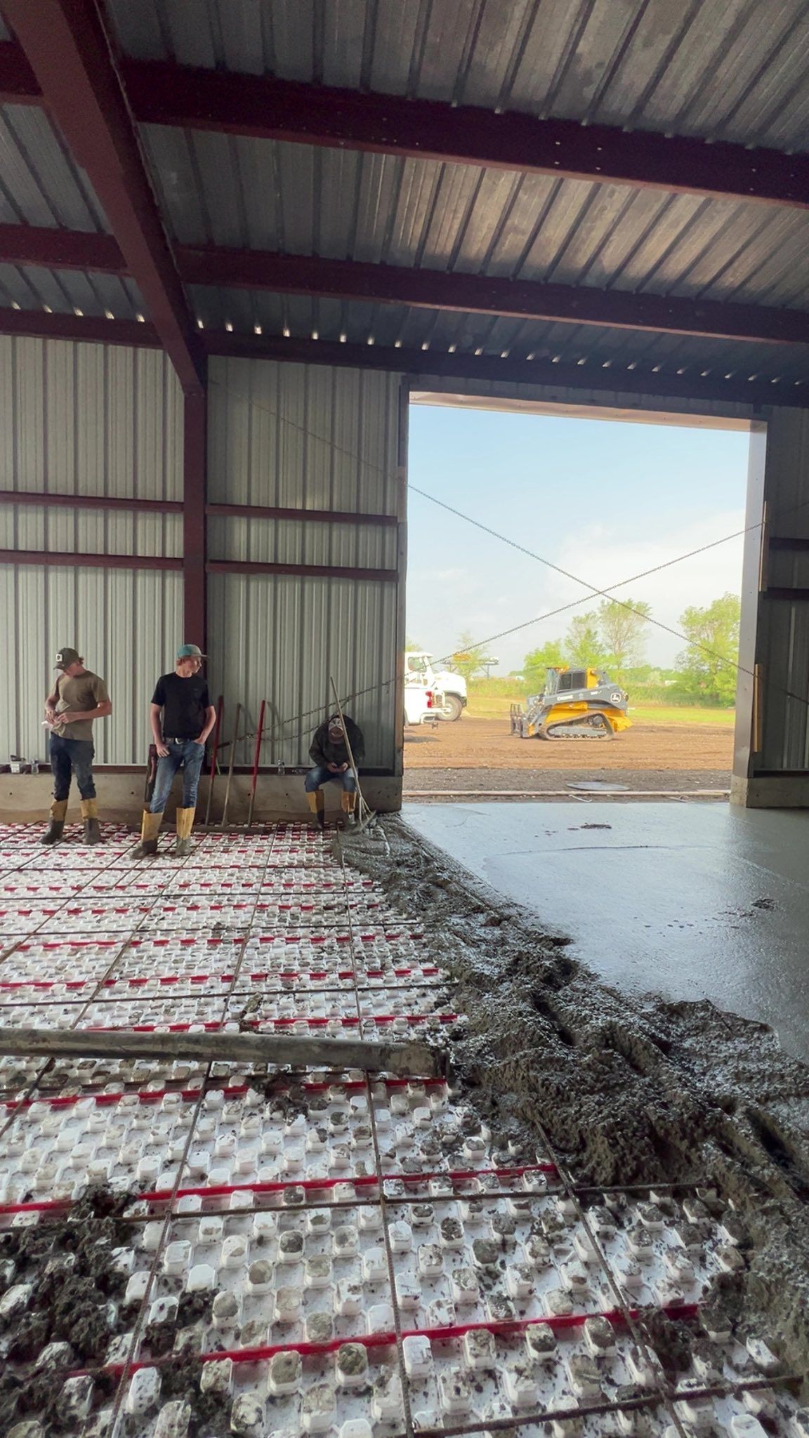 Construction workers pouring concrete inside a building with a large open doorway, other machinery visible outside.