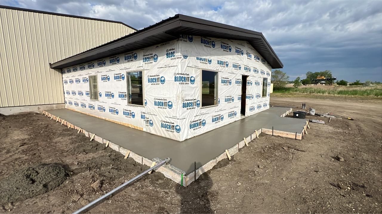 Construction site with a small building wrapped in blue sheathing, concrete foundation, and a gravel yard.