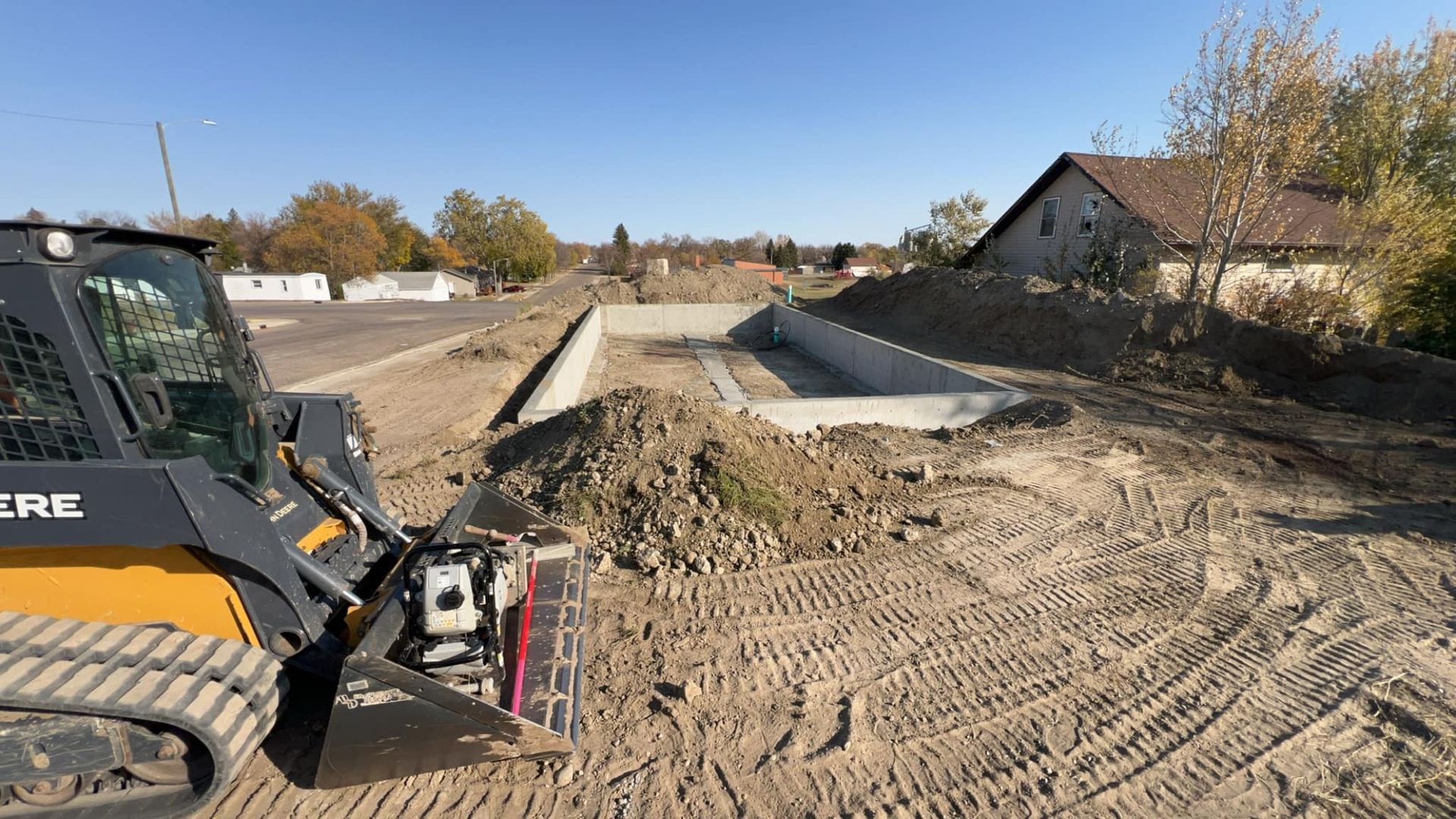 Construction site with a John Deere skid steer, dirt piles, and concrete foundations. Sunny day.