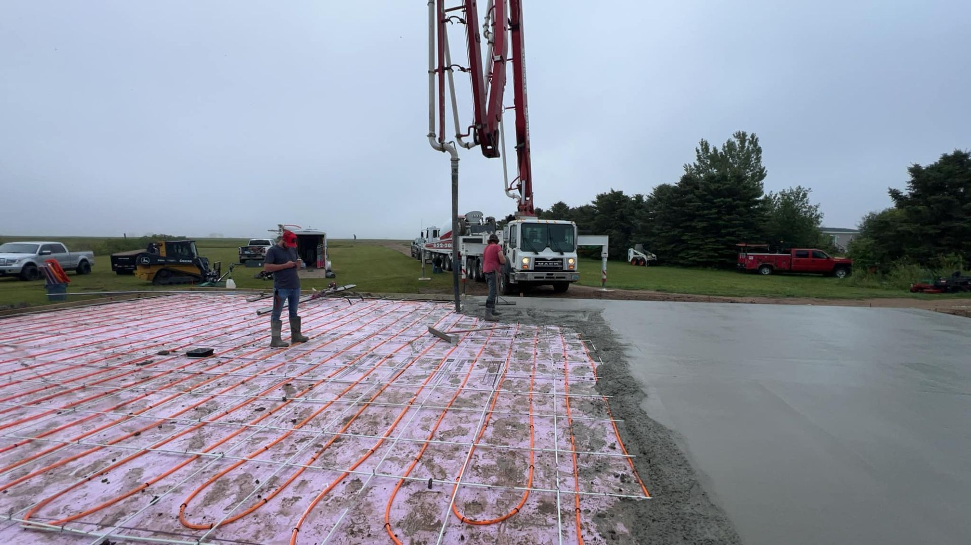 Concrete being poured over pink insulation with radiant heat tubing by a truck-mounted pump. Construction site.