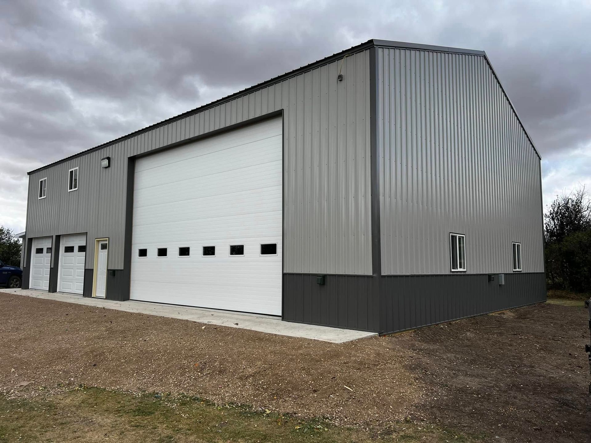 Large, grey metal building with three garage doors and small windows, on a gravel lot. Cloudy sky.