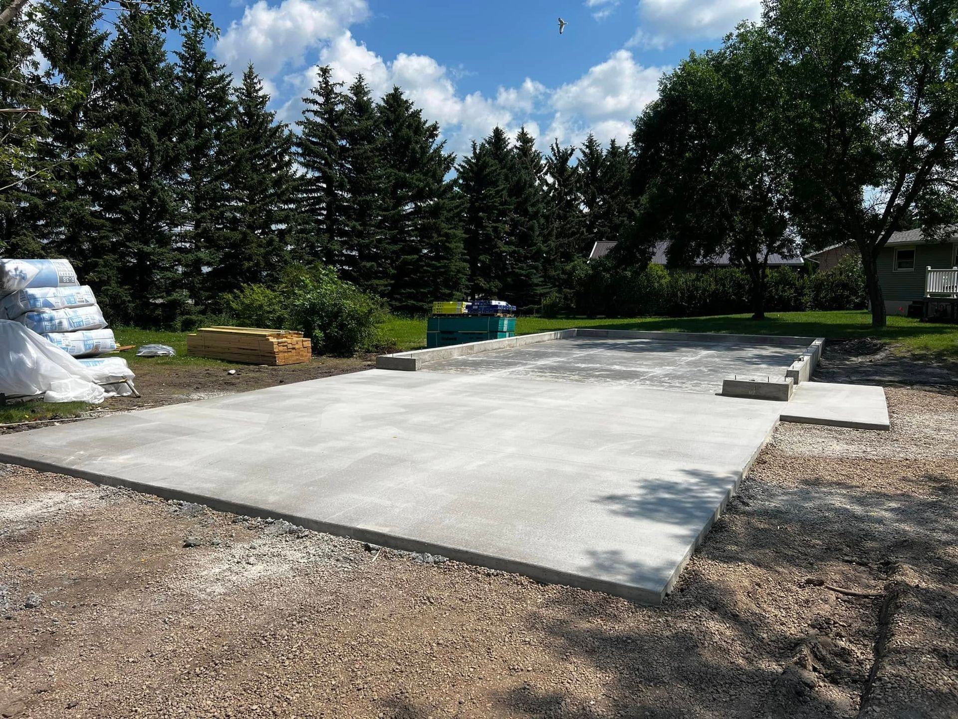 Newly poured concrete pad for a building, surrounded by gravel. Bags of materials and lumber are visible.