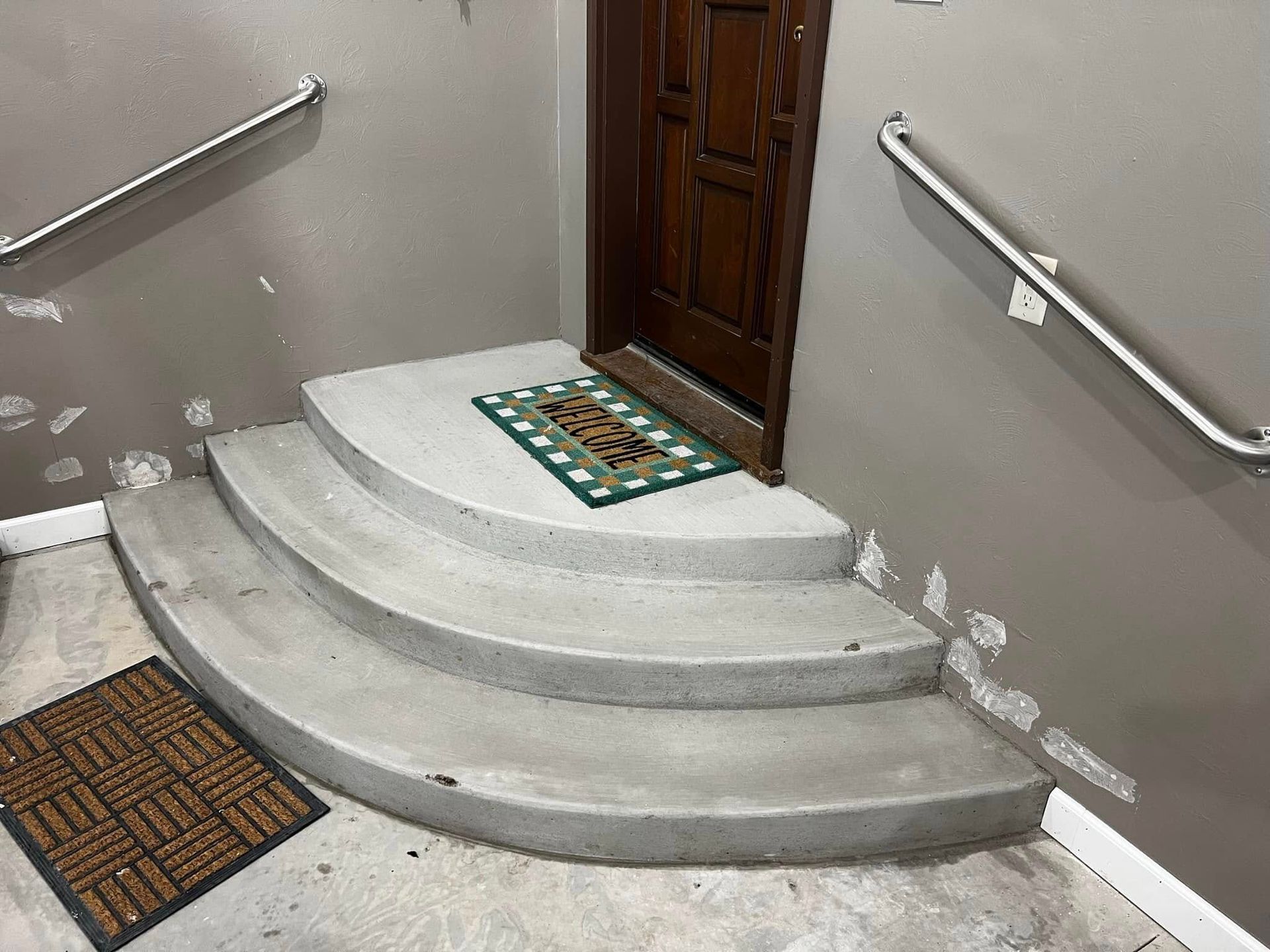 Concrete steps leading up to a wooden door, with handrails on either side. A doormat is on the landing.