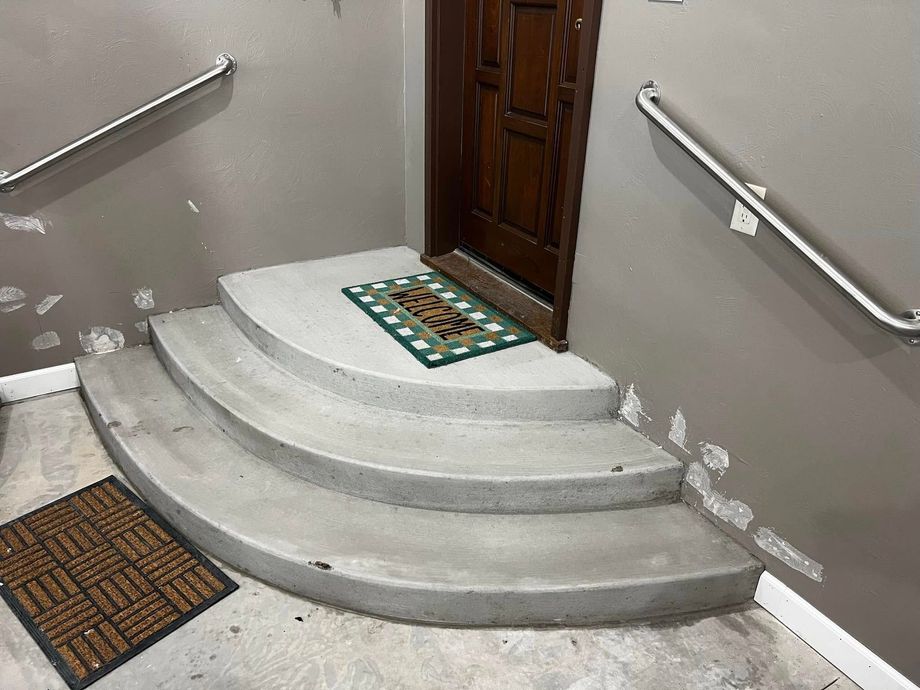 Concrete steps leading up to a wooden door with a welcome mat. Handrails on both sides, gray walls.