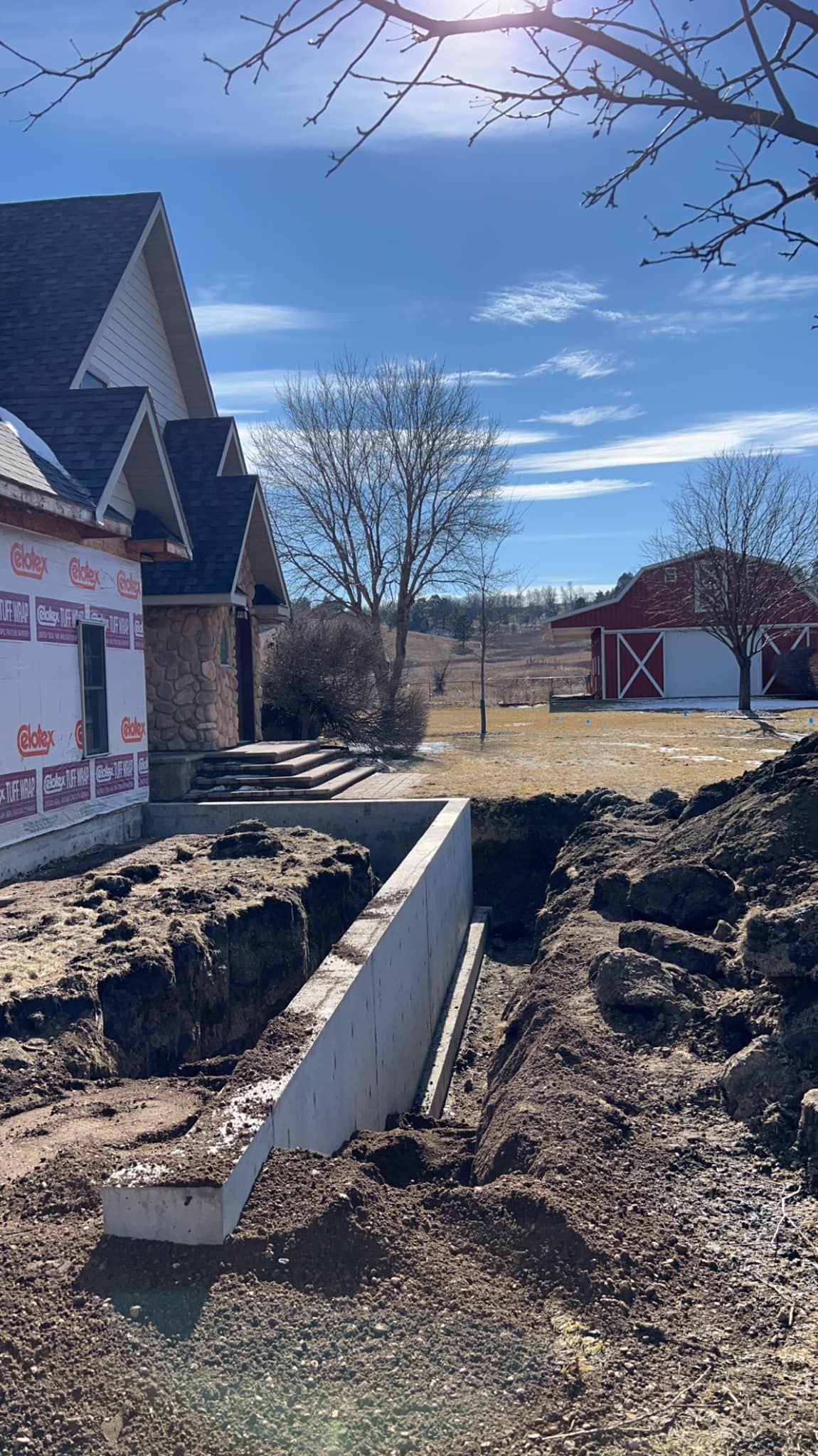 Construction site: house with exposed framing, concrete foundation, and red barn in background under a sunny, blue sky.