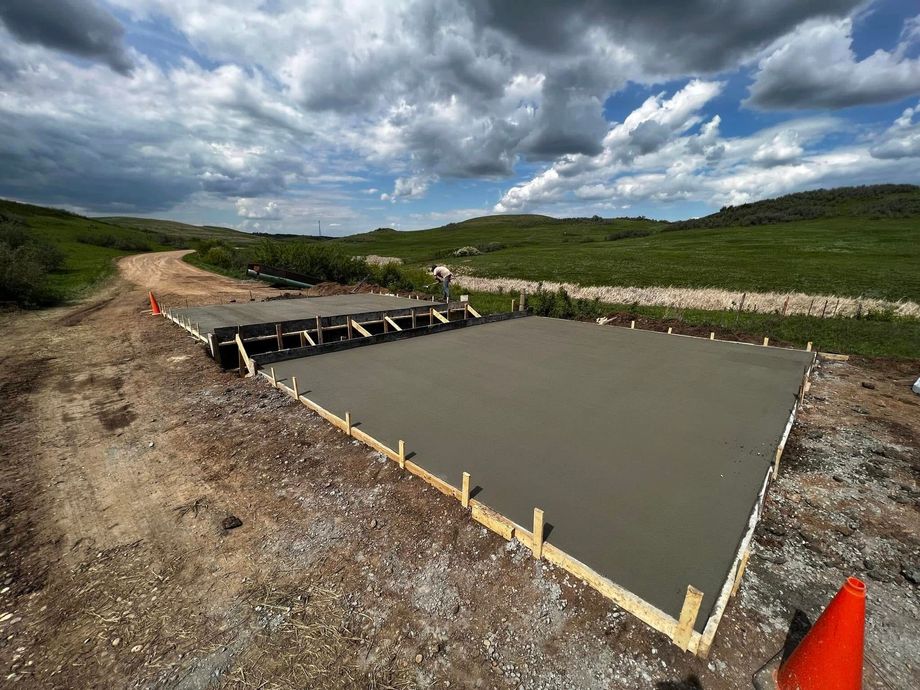 Freshly poured concrete slab with wooden formwork on a dirt road, under a cloudy sky.