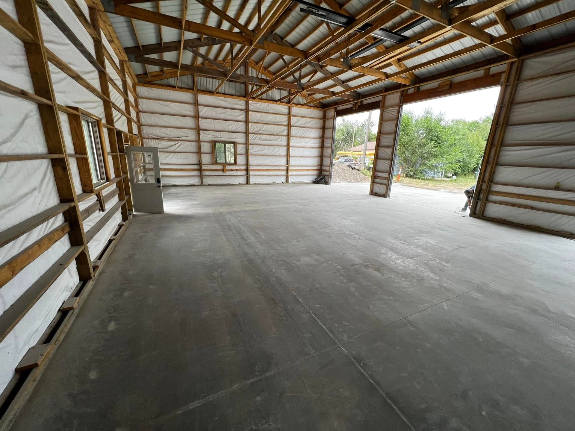 Empty concrete-floored building with wood framing and insulation. Open doorway leads to greenery.